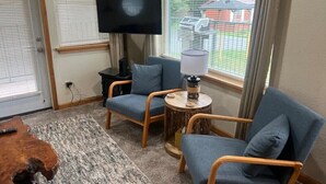Une pièce comprenant deux fauteuils gris, une petite table ronde avec une lampe, un téléviseur à écran plat fixé au mur et une table basse en bois.