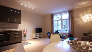 A modern kitchen with dark cabinets, a built-in oven, and a TV on a white stand. There is a dining area with a white table and chairs, and a large window with curtains. - The APARTMENTS Company - Bislett (Oslo)