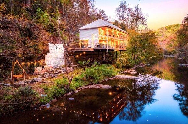Une maison à deux étages avec un balcon, située au bord d’une rivière paisible, entourée d’arbres.