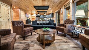 A cozy wooden interior with a central round coffee table, leather armchairs, and a kitchen area with modern appliances. - SwimmingHole RIVERFRONT Retreat - Fishing/HotTub/Firepit/Games/Private/NearGSMNP (Sevierville)