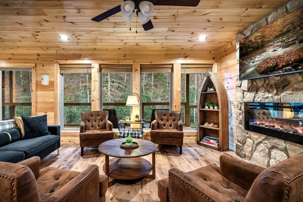 A wooden interior with a fireplace, leather chairs, a round table, and large windows. - RIVERFRONT Escape - Fishing/SwimmingHole/HotTub/Firepit/Games/Private/Near GSMNP (Sevierville)