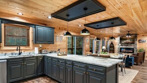 A spacious kitchen with dark cabinets, a large island, and a stone fireplace. - SwimmingHole RIVERFRONT Retreat - Fishing/HotTub/Firepit/Games/Private/NearGSMNP (Sevierville)