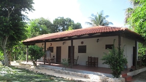 A single-story building with a tiled roof, a porch with chairs, and a small garden area with plants. - Casa de Toj2 (Chicxulub Pueblo)