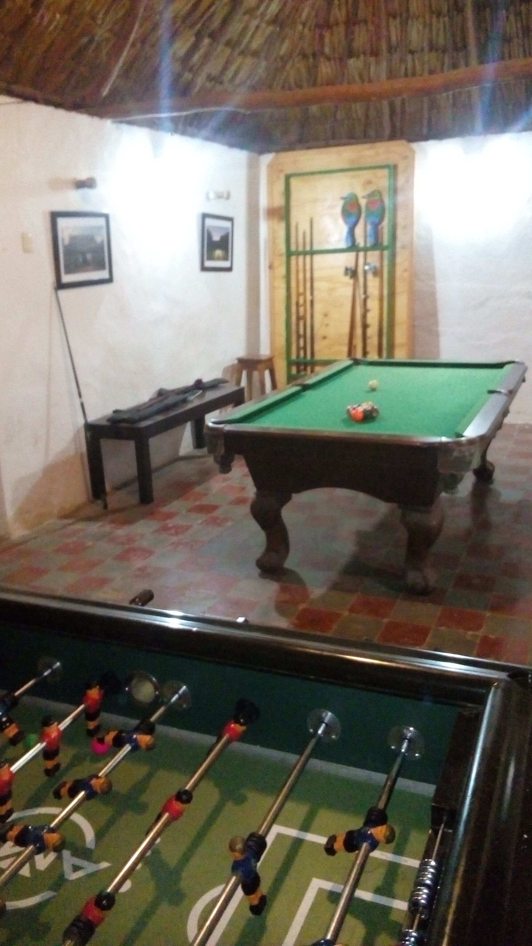 A pool table with a green felt surface, a foosball table in the foreground, and a wooden door with a green frame in the background.