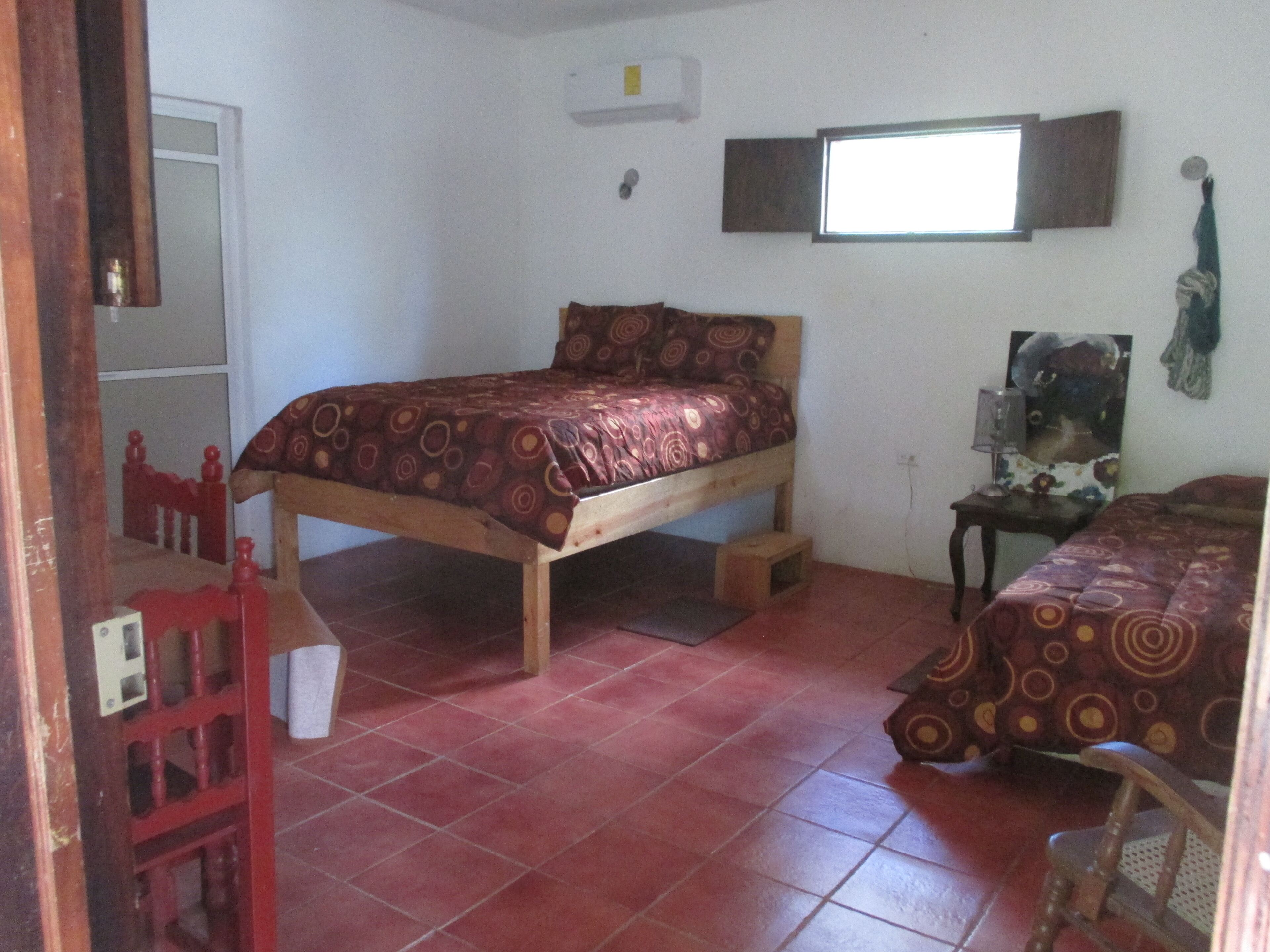 Una habitación pequeña con cama individual, estructura de madera, una silla roja y una mesita.