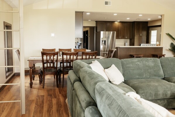 A modern living room with a green sofa, wooden dining table, and chairs. A kitchen with dark cabinets and stainless steel appliances is visible in the background.