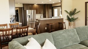 Une cuisine moderne avec des meubles en bois foncé, des appareils en acier inoxydable et un îlot central. On y trouve un coin repas avec des chaises et une table en bois, ainsi qu’un canapé vert au premier plan.