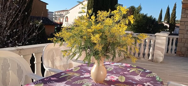 Una mesa con mantel floral, un jarrón con flores amarillas y sillas blancas en una terraza al aire libre con barandilla y edificios al fondo.