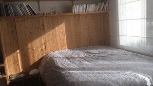 A bedroom with a wooden bunk bed, a small shelf with books, and a window with blinds.
