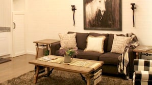 Ein gemütliches Wohnzimmer mit einem Holztisch, einem braunen Sofa und einem schwarz-weißen Schachbrettstuhl. Es befinden sich zwei Wandlampen und ein gerahmtes Gemälde eines Pferdes an der Wand.