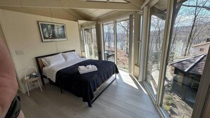 A bedroom with a large bed, a nightstand, and a window with a view of trees and a building. - The Escape Pod @Cheat Lake, private Hot Tub, near WVU (Morgantown)