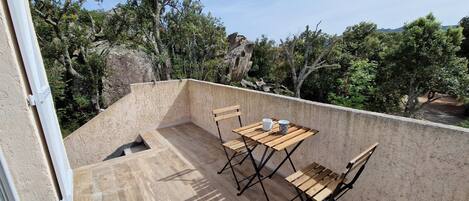 Ein Balkon mit Holztisch und -stühlen, einem kleinen Fenster und Blick auf Bäume und Hügel.
