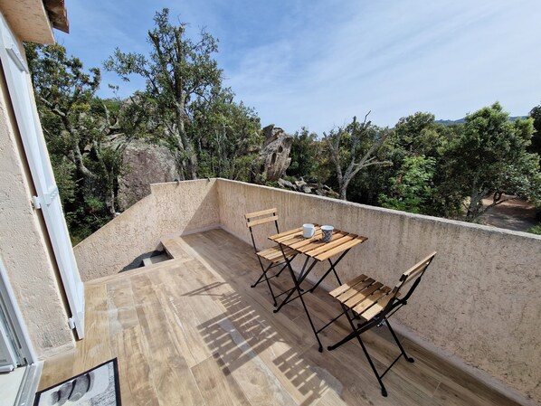 A balcony with a wooden table and chairs, a small window, and a view of trees and hills. - Appartement 27 M2 Baie DE Santa Giulia Ideal Pour 2 Personnes (Porto-Vecchio)
