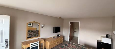 Un dormitorio con cama, escritorio, silla, cómoda, televisor y una puerta que da a otra habitación.