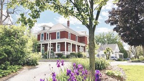 A two-story brick house with a front porch, surrounded by greenery and purple flowers.