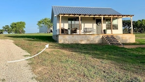 A small, single-story house with a porch and stone foundation, surrounded by a grassy field and trees. - The Ranch@Fredericksburg - Magnolia Cottage - 1BD/1BA (Fredericksburg)