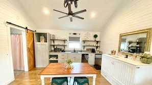 A kitchen with a wooden table, two green stools, white cabinetry, and a ceiling fan. - The Ranch@Fredericksburg - Magnolia Cottage - 1BD/1BA (Fredericksburg)
