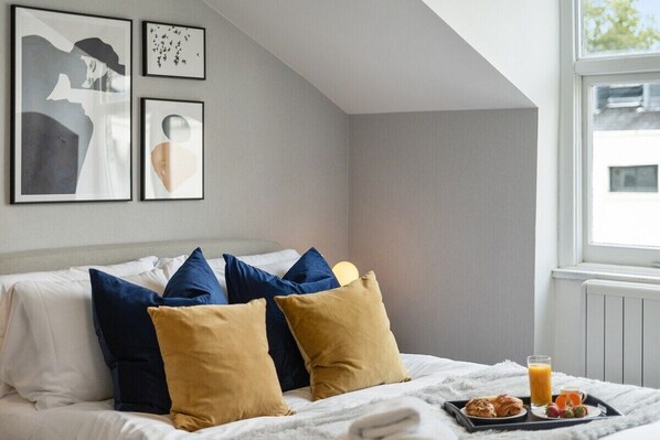 Una cama bien hecha con almohadas azules y mostaza, una bandeja con cruasanes y fruta, y un vaso de jugo de naranja.