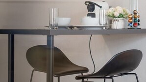 A modern kitchen with a glass-top table, two black chairs, a coffee machine, and a shelf with jars and a flower vase. - Stylish Studio with Private Bath • Near Hyde Park (Greater London)