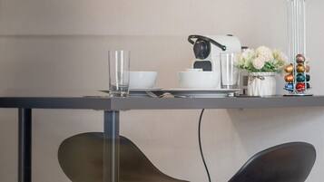 A modern kitchen with a glass-top table, two black chairs, a coffee machine, and a shelf with jars and a flower vase.
