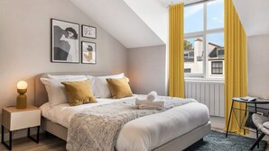 A modern bedroom with a large bed, a bedside table with a lamp, and a window with yellow curtains. - Stylish Studio with Private Bath • Near Hyde Park (Greater London)