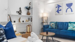 A modern living room with a blue sofa, a round coffee table, and a white armchair. There are decorative pillows, a vase with plants, and a lamp on a side table.