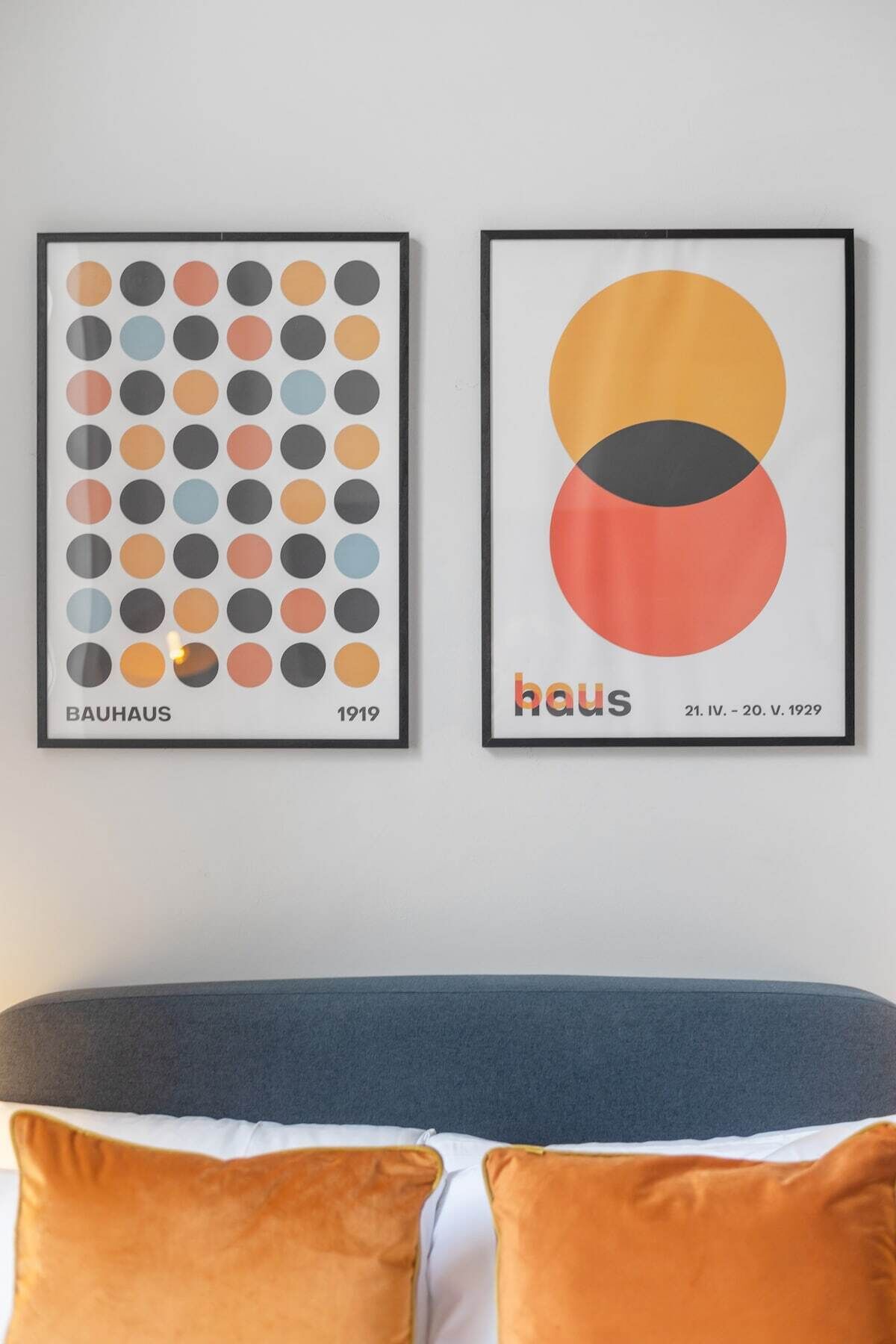 Zwei gerahmte Kunstwerke an einer Wand über einem Bett mit grauem Kopfteil und orangefarbenen Kissen.