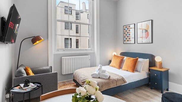 Ein modernes Hotelzimmer mit großem Fenster, einem Bett mit blauer Kopfteil, einem Nachttisch mit Lampe, einem grauen Sofa und einem kleinen Tisch mit Zeitschrift und Dekorationsobjekt.