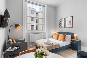 Habitación de hotel moderna con un ventanal grande, una cama con cabecera azul, una mesita de noche con lámpara, un sofá gris y una pequeña mesa con una revista y un objeto decorativo.