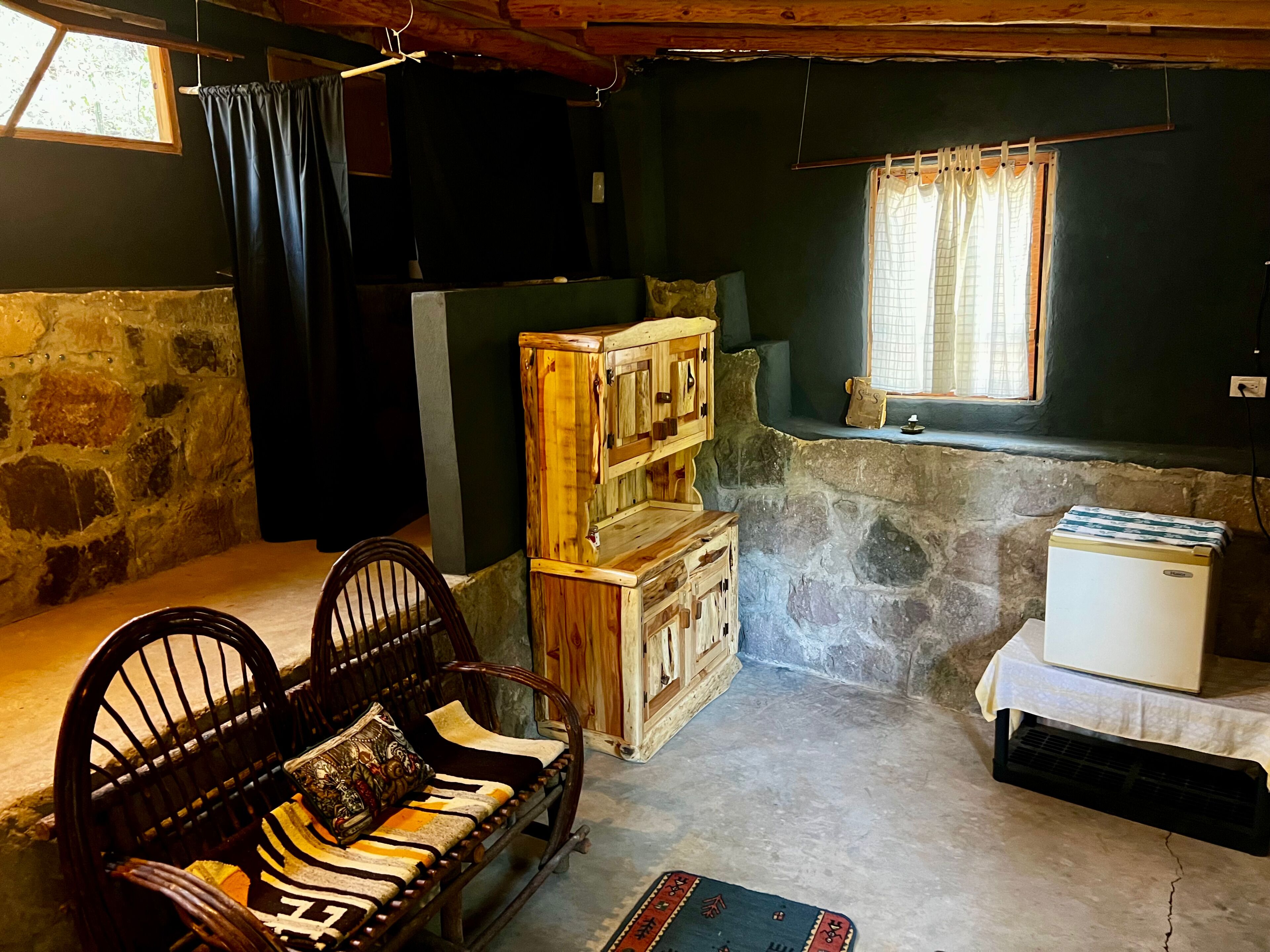 Una habitación rústica con pared de piedra, muebles de madera, un banco, una mesita y una ventana con cortinas.
