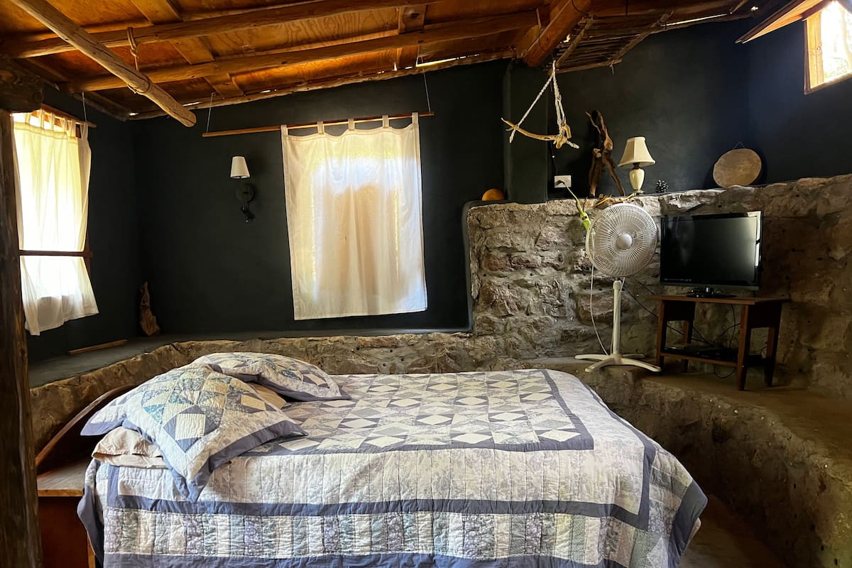 A rustic room with a bed, a stone wall, a television, and a fan.