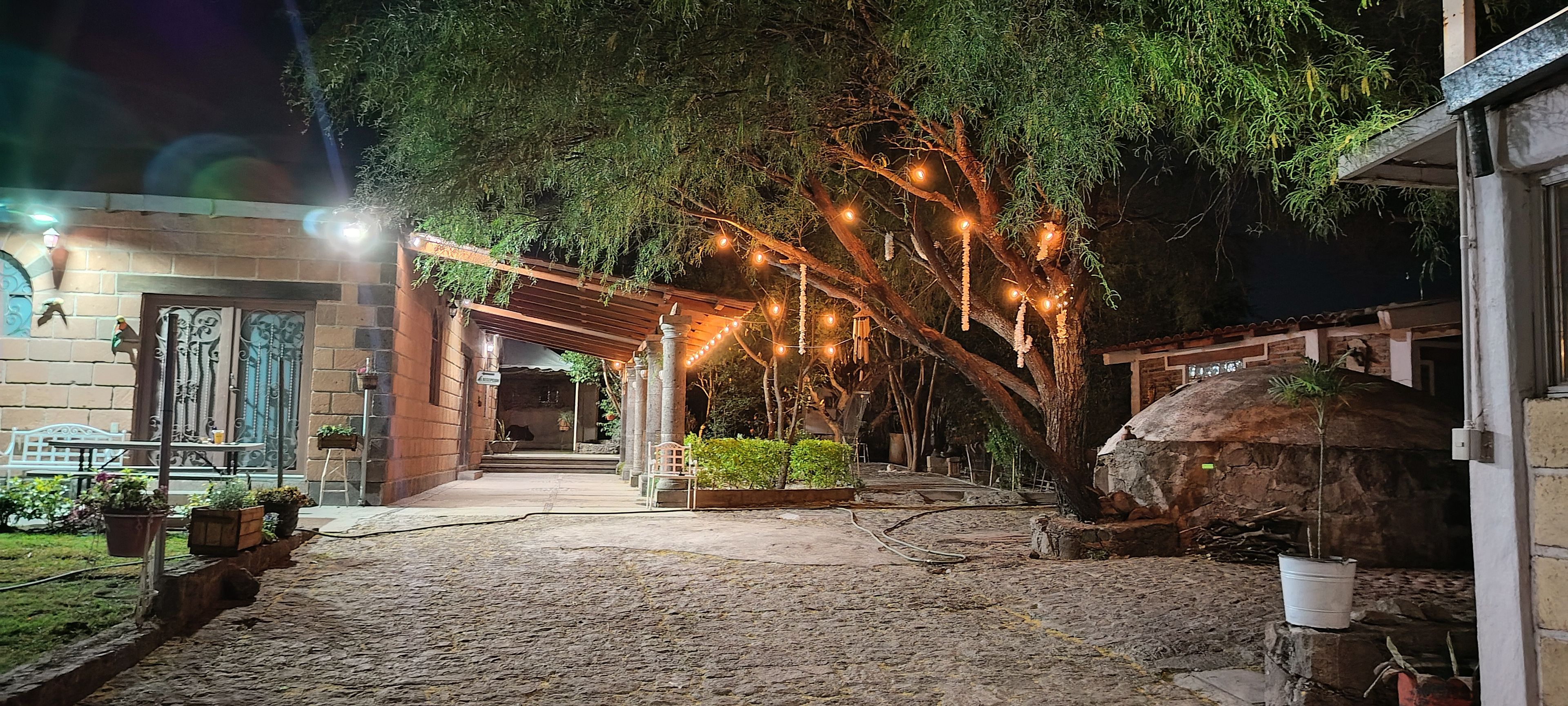Ein Innenhof mit einem gepflasterten Weg, einer Pergola mit Lichterketten und einem rustikalen Backofen aus Stein.