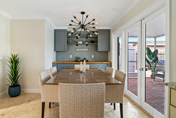 A dining area with a large wooden table and patterned chairs, a ceiling light fixture, and a kitchen area with a 'Coffee Bar' sign.
