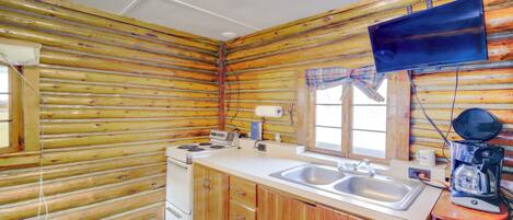A log cabin interior with a kitchen area featuring wooden cabinets, a stove, a sink, and a mounted television.