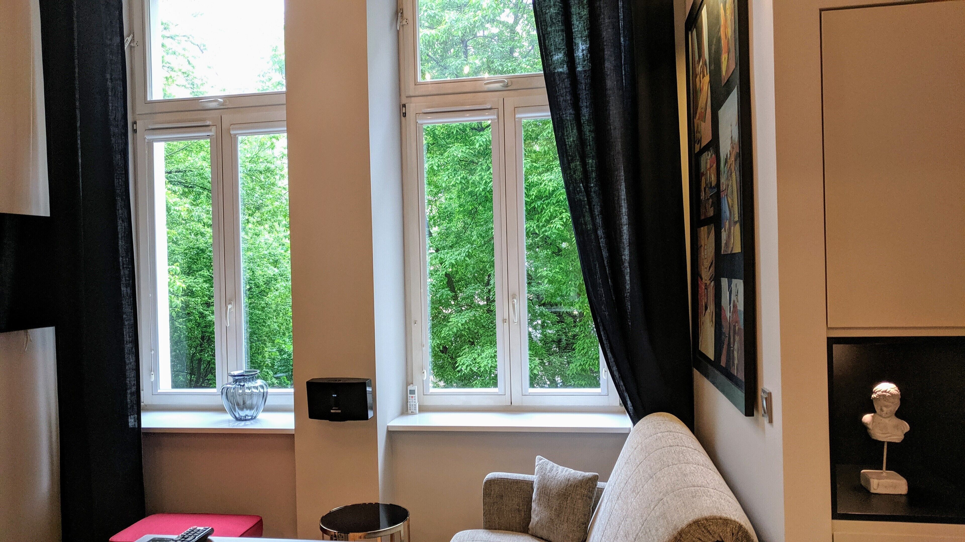 Een kamer met twee ramen, een bank, een kleine tafel en een wandplank met een borstbeeld.