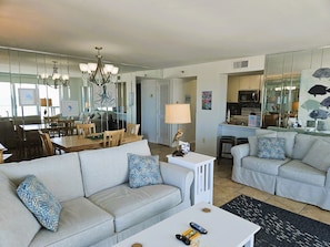 A modern living room with two grey sofas, a dining area with wooden furniture, and a kitchen visible through an open door. - Panoramic 10th Floor Gulf & Bay Views, Pool, Tennis Court- TT10B (Pensacola Beach)