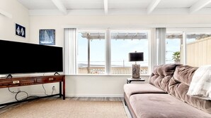 A living room with a large flat-screen TV, a wooden coffee table, a beige sofa, and a view of the ocean through large windows.