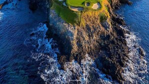 A coastal golf course with a water body, rocky terrain, and a distant residential area.