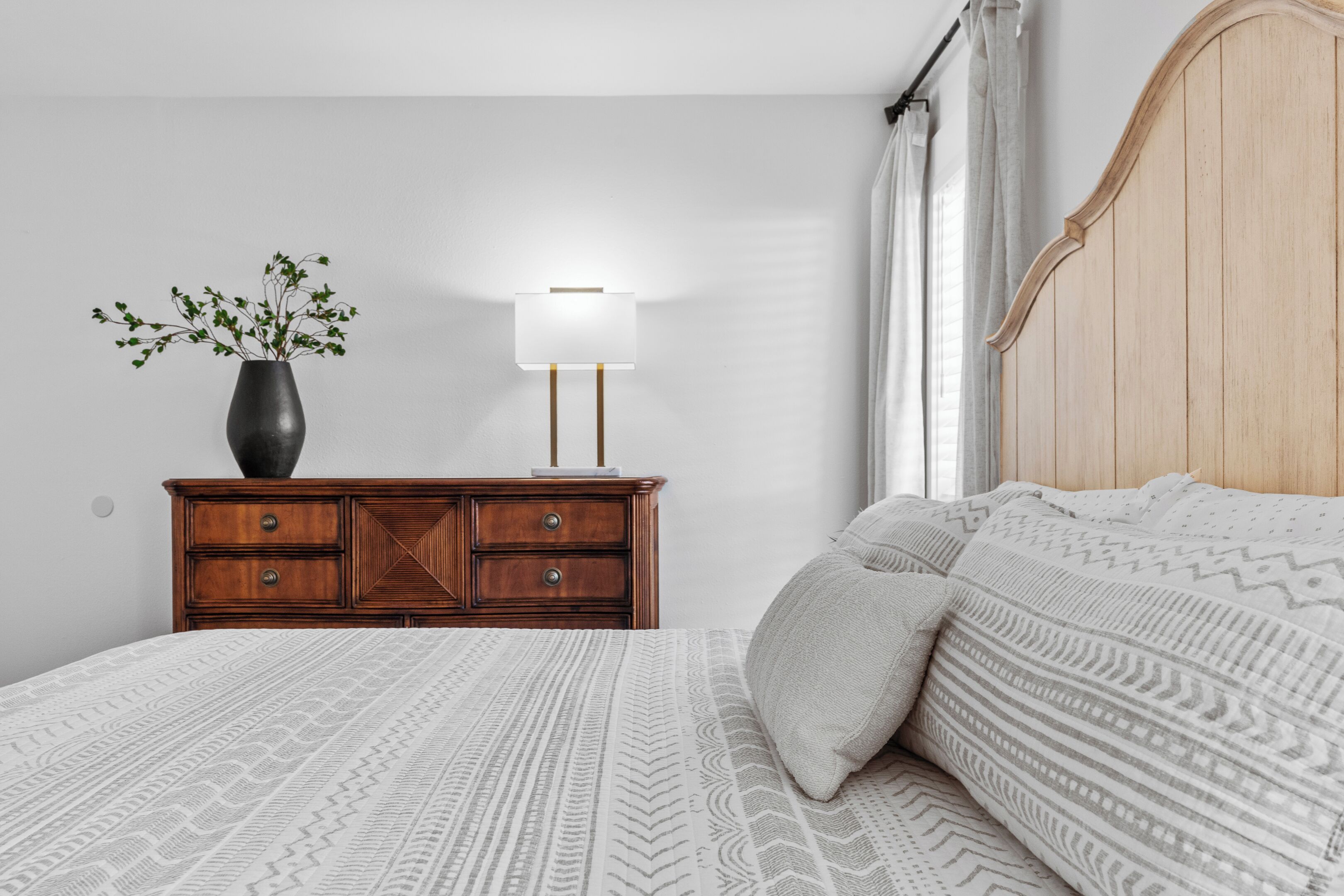 Ein ordentlich bezogenes Bett mit gemusterten Betttextilien, eine hölzerne Kommode mit Schubladen, eine Vase mit kleinen Pflanzen und eine Nachttischlampe.