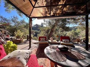 Un área de estar al aire libre con vistas a una ladera boscosa. Hay dos sillones acolchados y una mesita con un cuenco y un objeto decorativo.