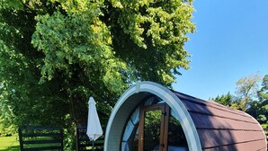 A small, semi-circular structure with a glass door and windows, surrounded by gravel, with a bench and umbrella nearby. - Priory Glamping Pods (Killarney)