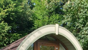 A small, semi-circular, wooden structure with a door, surrounded by greenery and a red object on the ground. - Priory Glamping Pods (Killarney)