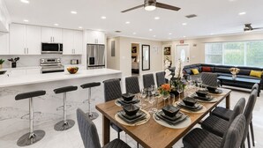 A modern kitchen with white cabinets, a microwave, and a refrigerator. There is a dining area with a wooden table set for six, surrounded by chairs.