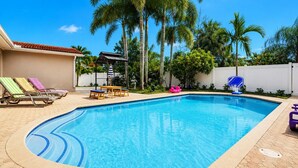 A swimming pool with lounge chairs and a shaded area, surrounded by palm trees and a white fence.