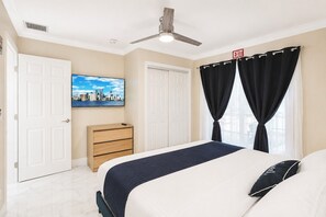 Una habitación de hotel con cama, cómoda, ventilador de techo, televisión y ventana con cortinas.