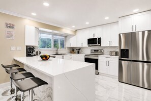 Cocina moderna con armarios blancos, electrodomésticos de acero inoxidable, una isla grande con taburetes y una ventana con persianas.