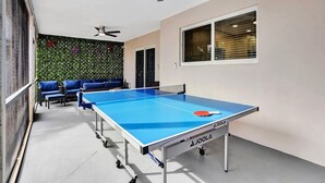 A ping pong table with a blue playing surface and a red paddle, set in a room with a ceiling fan, a window with blinds, and a green leafy wall.