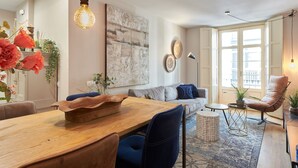 A modern living room with a wooden dining table, blue chairs, a grey sofa, and a large abstract painting on the wall.