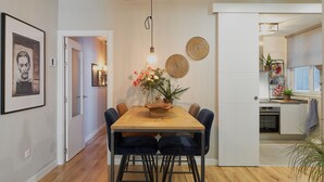 A dining area with a wooden table, six chairs, and a vase of flowers. There is a kitchen visible in the background with a window and a plant on the counter.