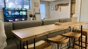 Uma sala de estar moderna com uma longa mesa de madeira, quatro banquetas, um sofá cinza e uma televisão exibindo diversos canais.